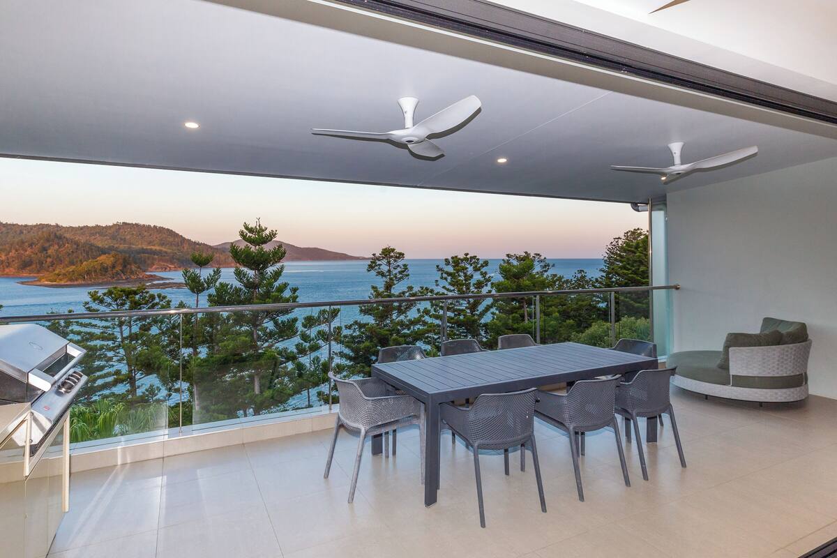 A balcony with a table and chairs, a grill, and a view of the sea and hills.