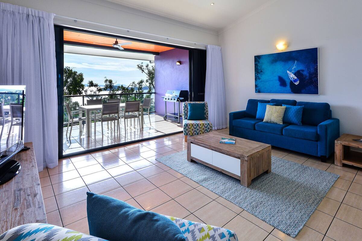 A modern living room with a blue sofa, a coffee table, and a large window overlooking a patio with outdoor furniture.