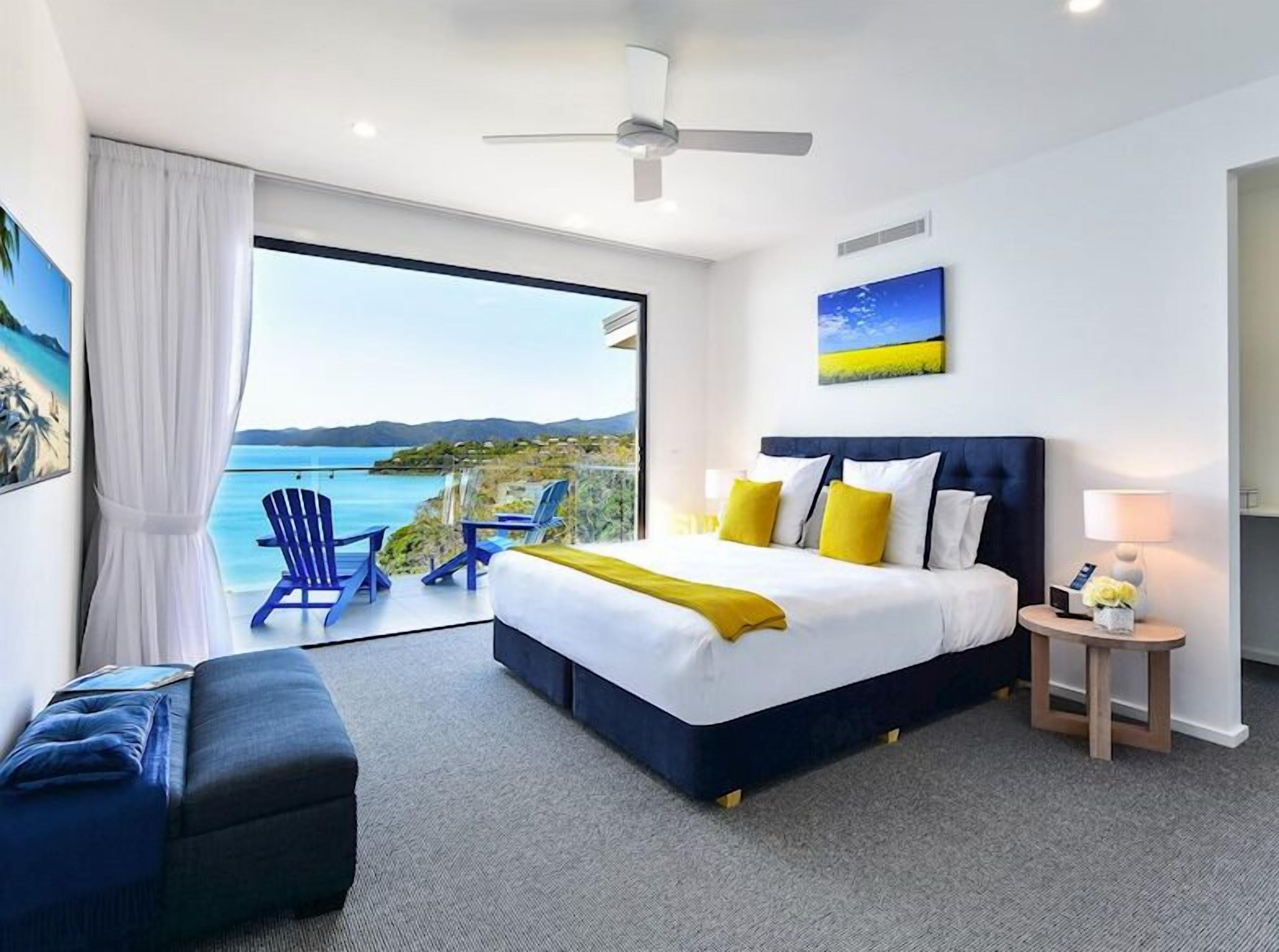 A modern hotel room with a large bed, a ceiling fan, a view of the sea, and a balcony with blue chairs.