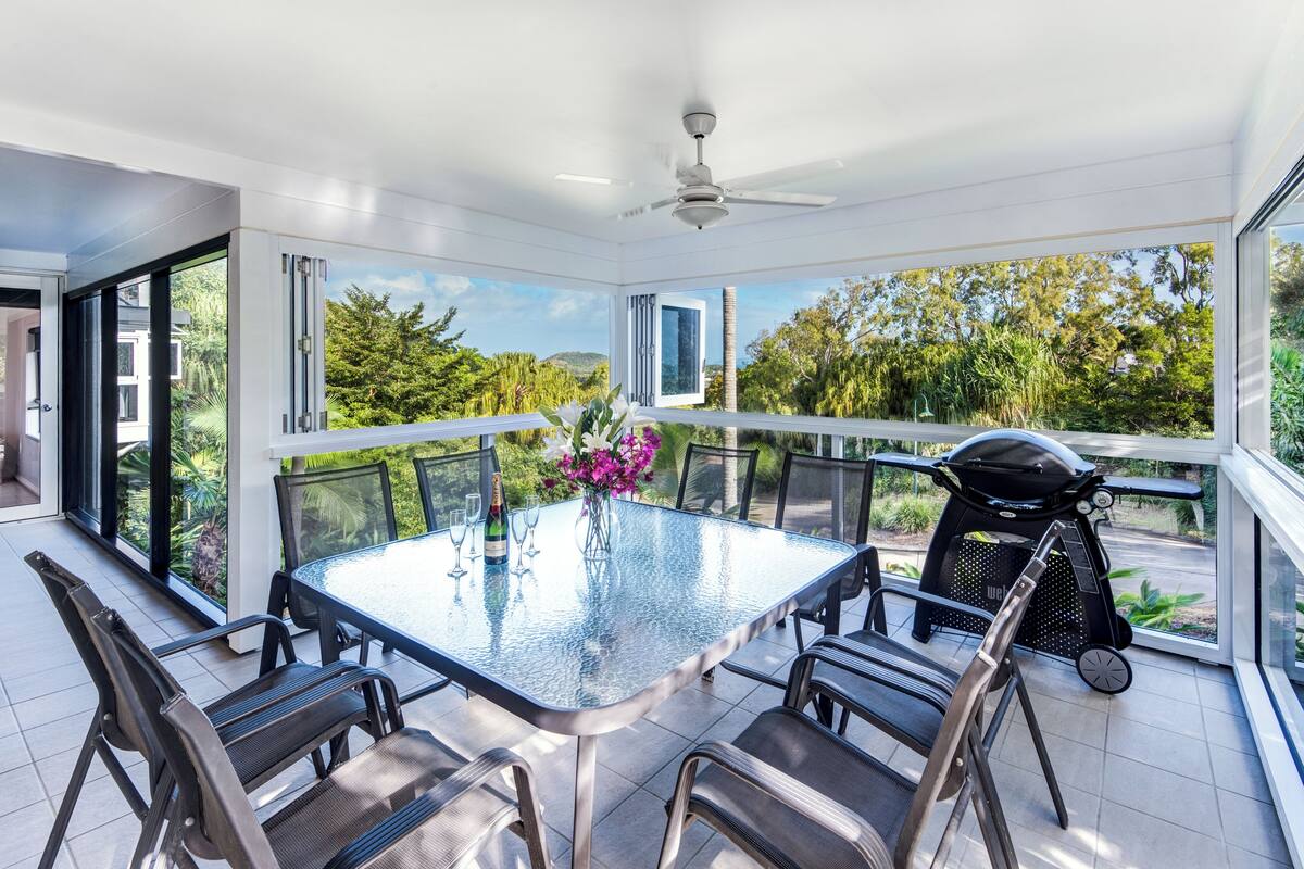A sunlit room with a glass table, chairs, a ceiling fan, and a view of greenery.