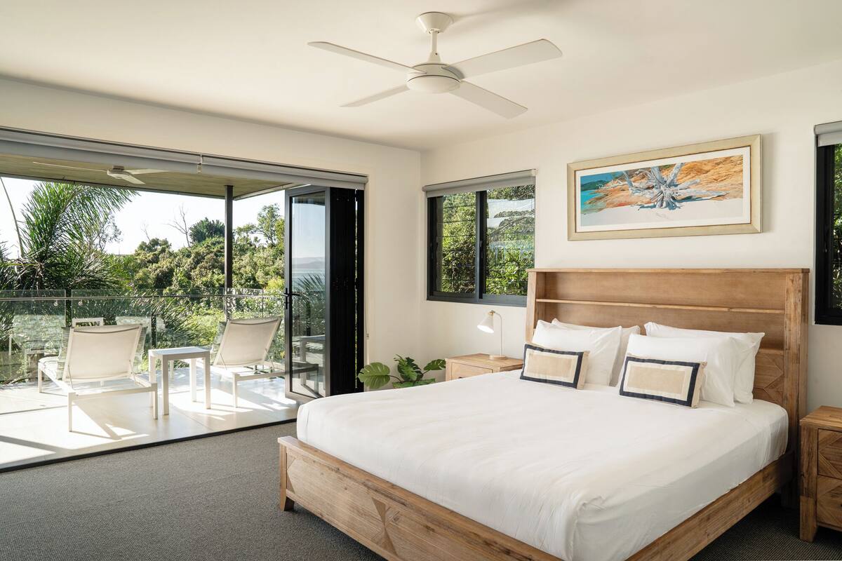 A bedroom with a large bed, a ceiling fan, a painting, and a view of greenery outside.