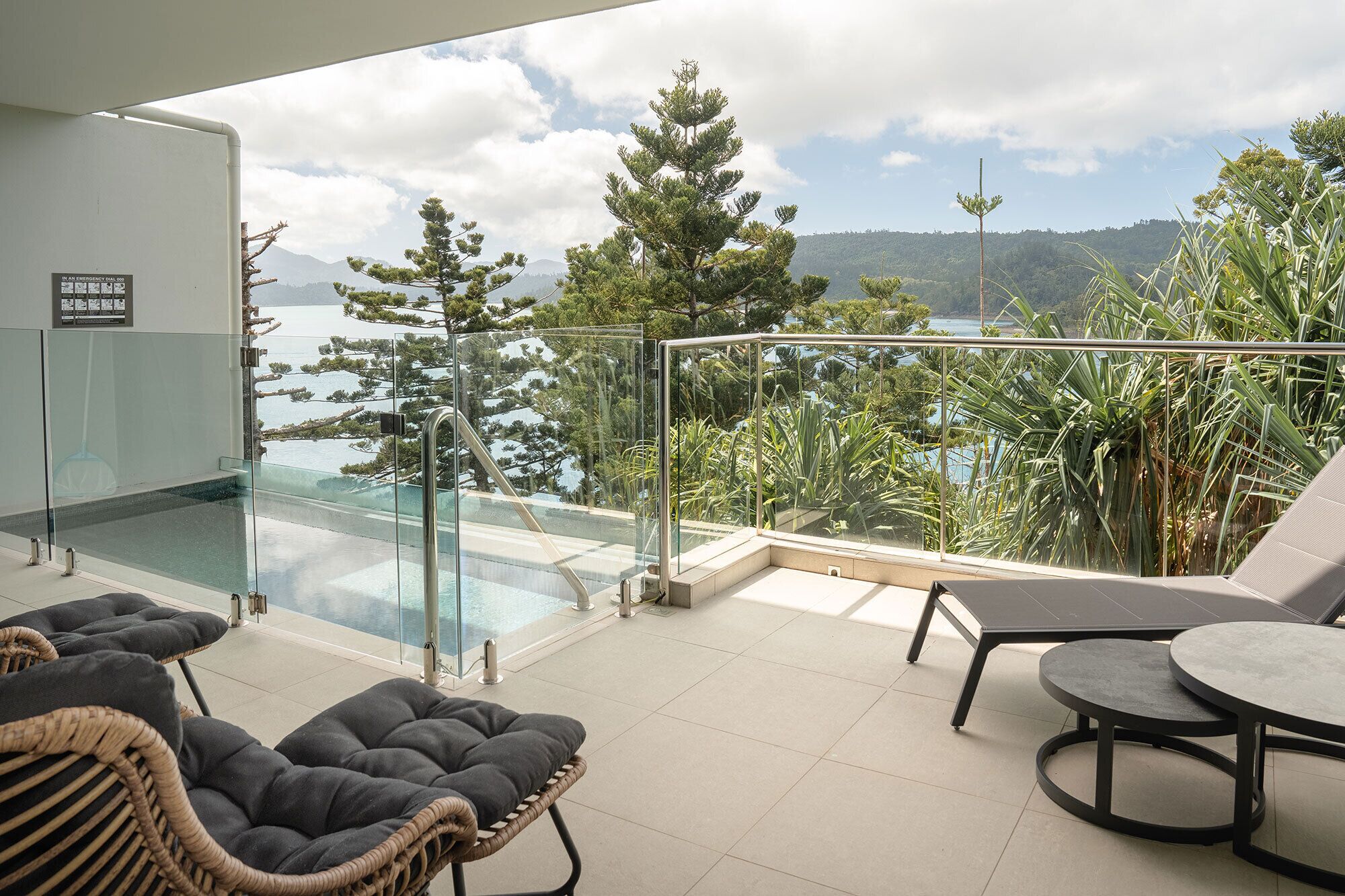 Un balcon doté d’une balustrade en verre, une piscine, des fauteuils en osier, une table et une vue sur la mer et les arbres.