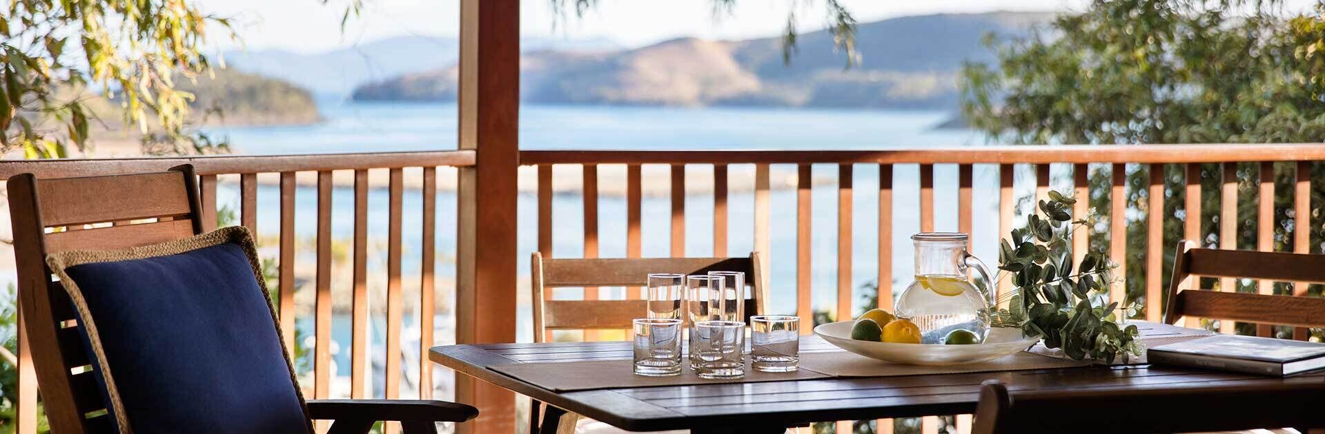 A wooden deck with a table set for two, featuring a pitcher, glasses, and a bowl of fruit.