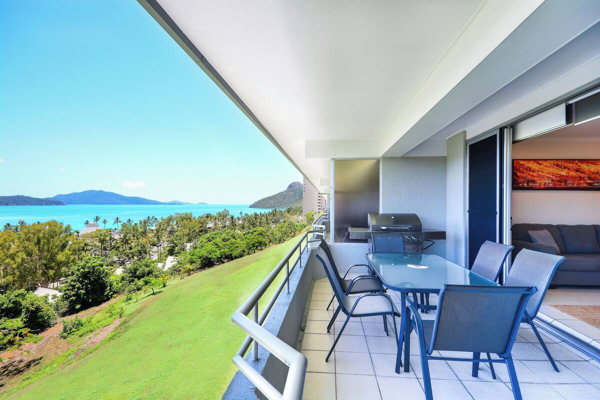 A balcony with a glass table and chairs, a grill, and a view of the ocean and hills.