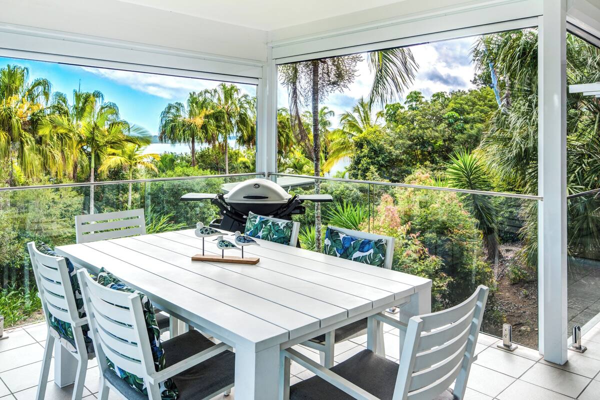 A dining area with a white table and chairs, a grill, and a view of lush greenery and palm trees.