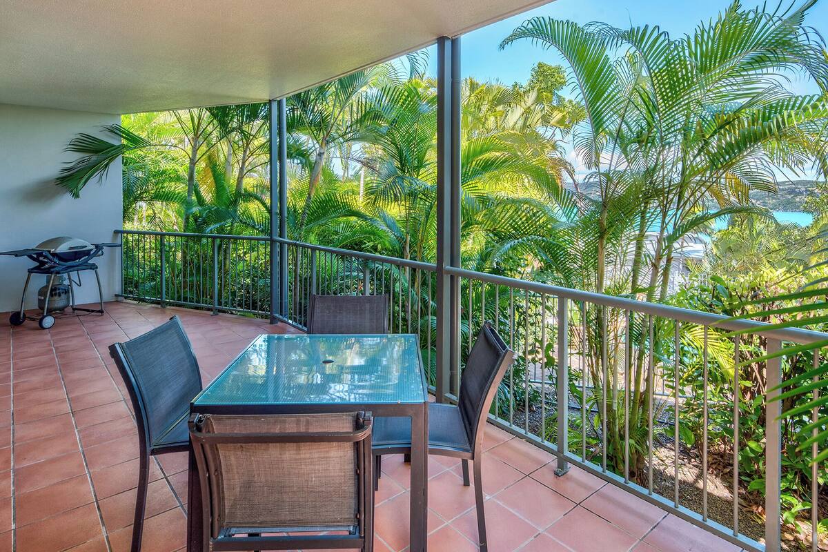 A balcony with a glass table and chairs, surrounded by metal railings and lush tropical vegetation.