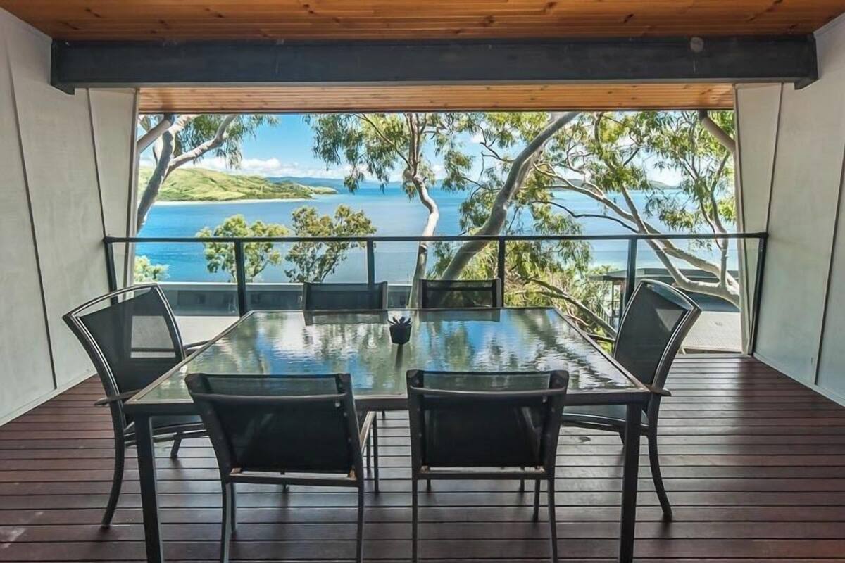 A deck with a glass table and chairs, overlooking a scenic view of the sea and greenery.