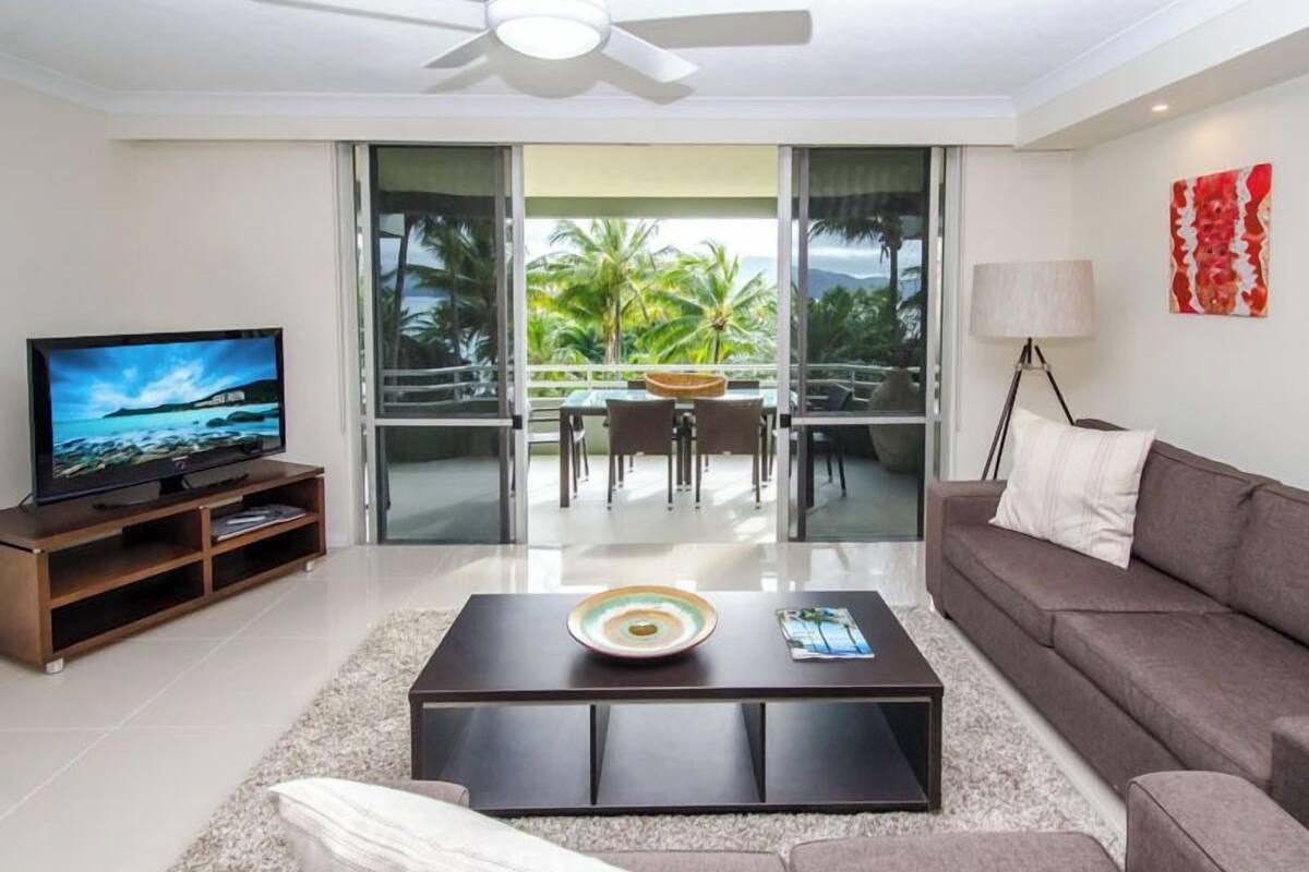 A modern living room with a flat-screen TV, a sofa, a coffee table, and a balcony with a view of palm trees.