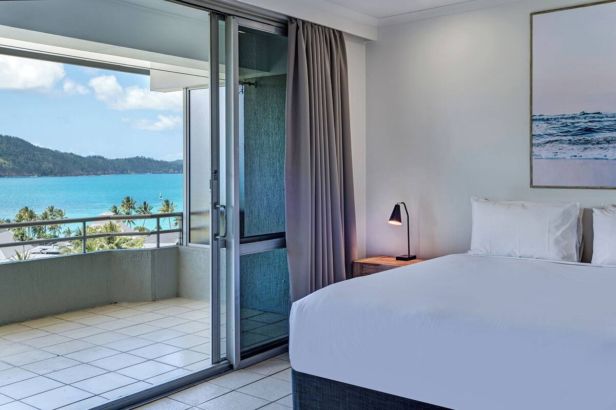 A hotel room with a bed, a nightstand, a lamp, a sliding glass door leading to a balcony with a view of the sea and hills.