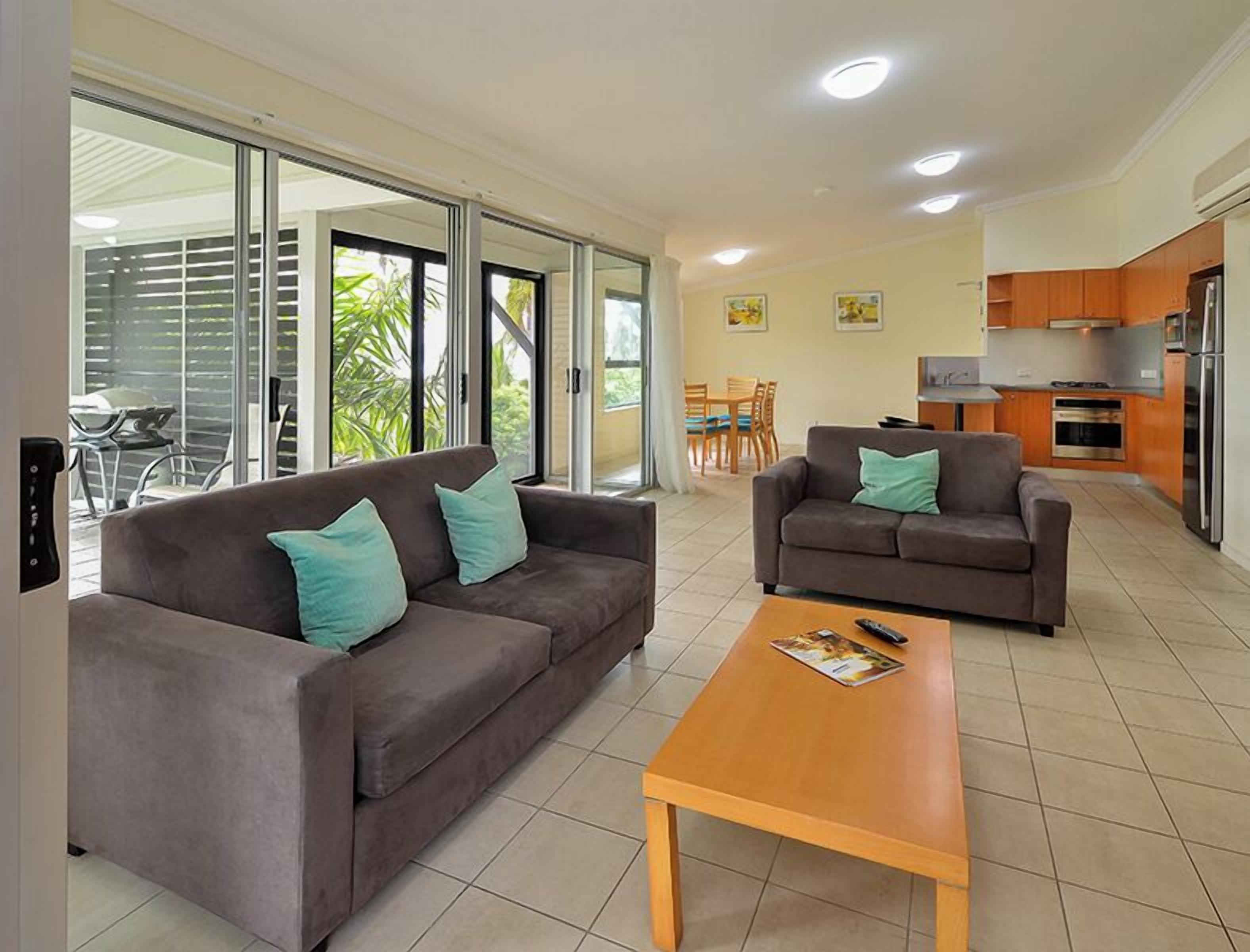 A modern living room with two sofas and a coffee table, a kitchen area with appliances, and a sliding glass door leading to a patio with outdoor furniture.
