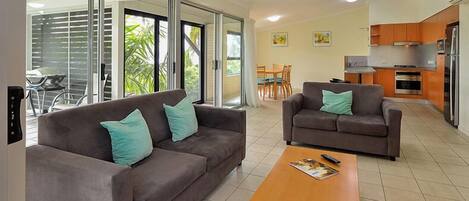 A modern living room with two sofas and a coffee table, a kitchen area with appliances, and a sliding glass door leading to a patio with outdoor furniture.