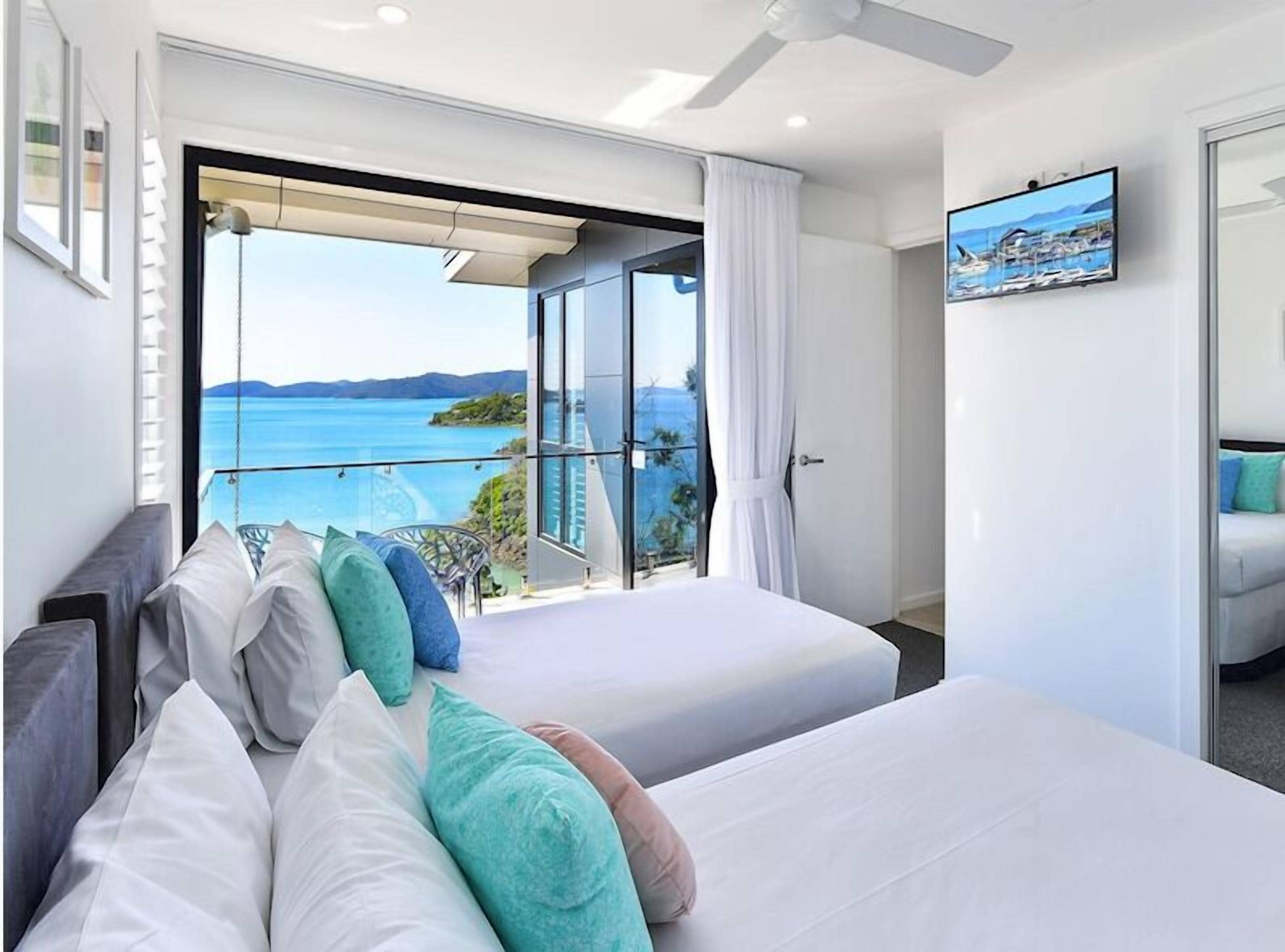 A hotel room with a large balcony, a bed with multiple pillows, a TV, and a view of the sea.