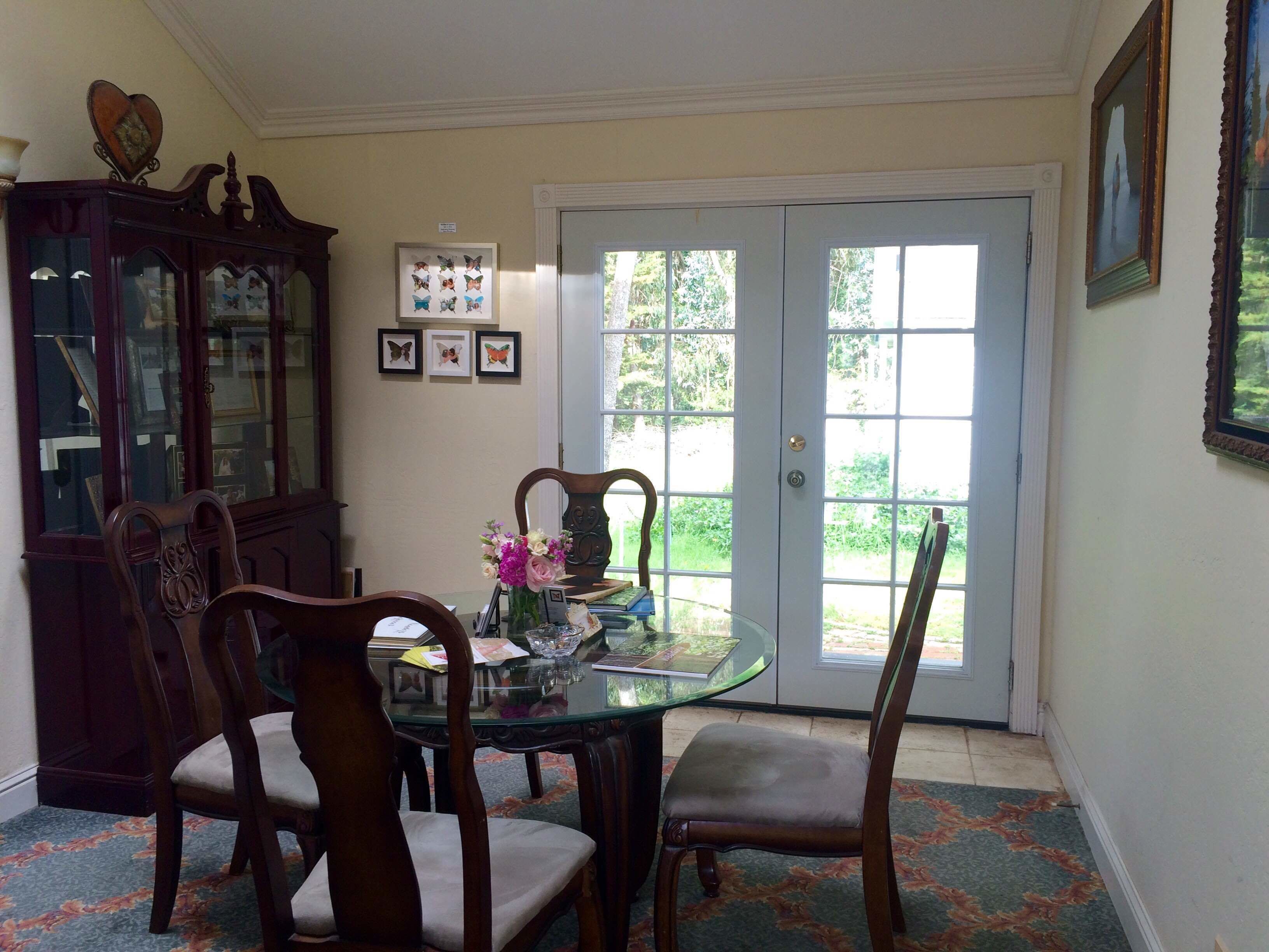 Uma sala de jantar com mesa de vidro cercada por cadeiras de madeira, um arranjo floral no centro e vista através de uma porta de vidro para uma área externa.