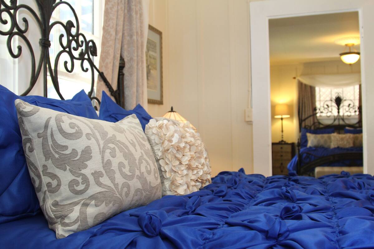 A bed with blue bedding and patterned pillows, a black metal headboard, and a view of a room with a lamp and another bed in the background.