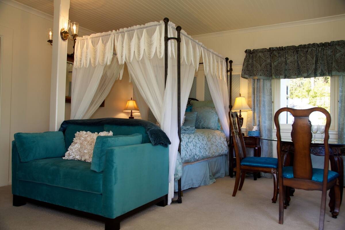 A bedroom with a four-poster bed, a teal sofa, a wooden chair, and a desk with a lamp.
