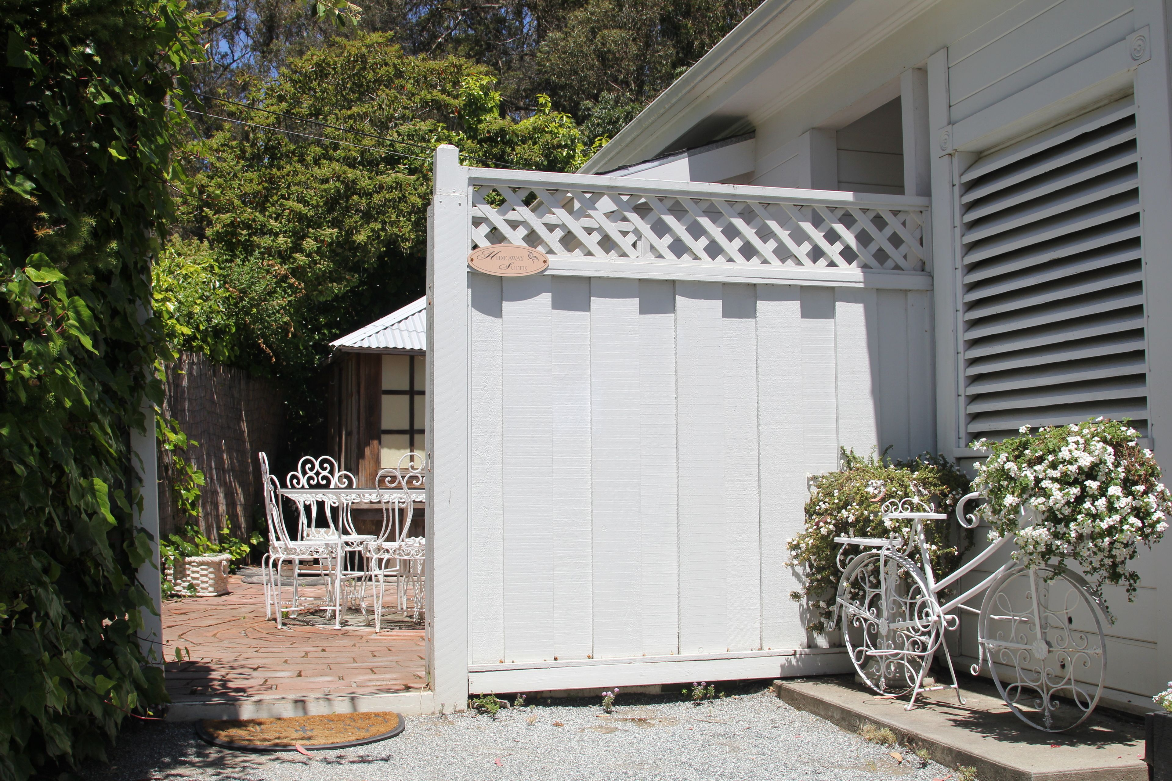 A white fence with a lattice top, a white bicycle with a flower basket, and a patio with white metal chairs and a table.