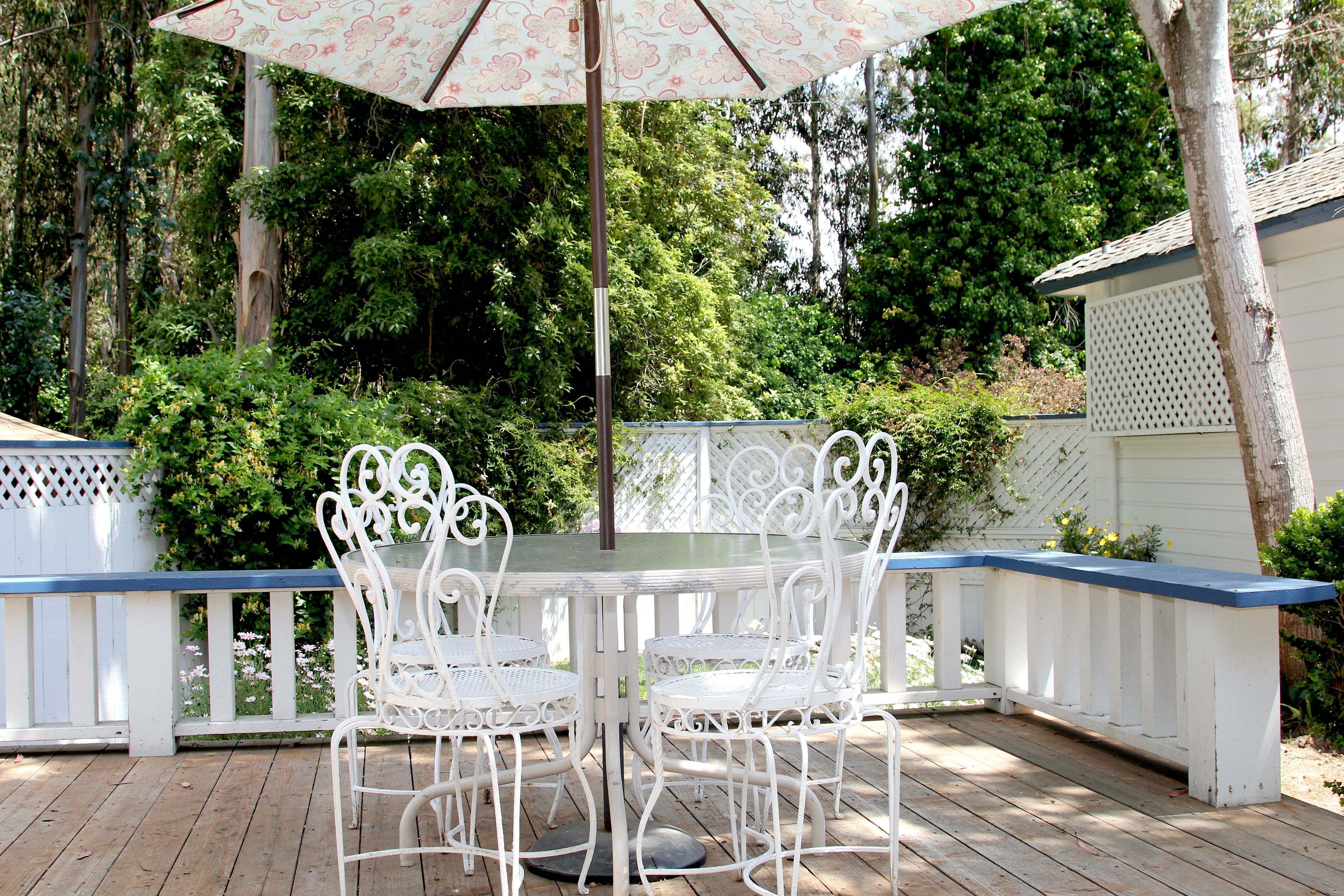 A white outdoor patio with a glass table and chairs, surrounded by a white fence and lush greenery.