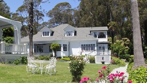 A two-story house with a white exterior, a front porch with white railings, and a small garden with pink flowers and a white table with chairs.