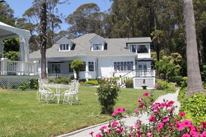 A two-story house with a white exterior, a front porch with white railings, and a small garden with pink flowers and a white table with chairs.
