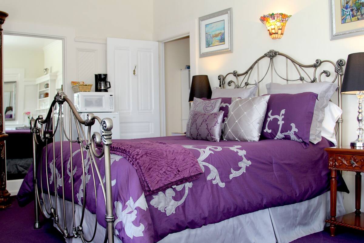 A bedroom with a bed, a nightstand, a lamp, a mirror, a door, and a wall-mounted light fixture.