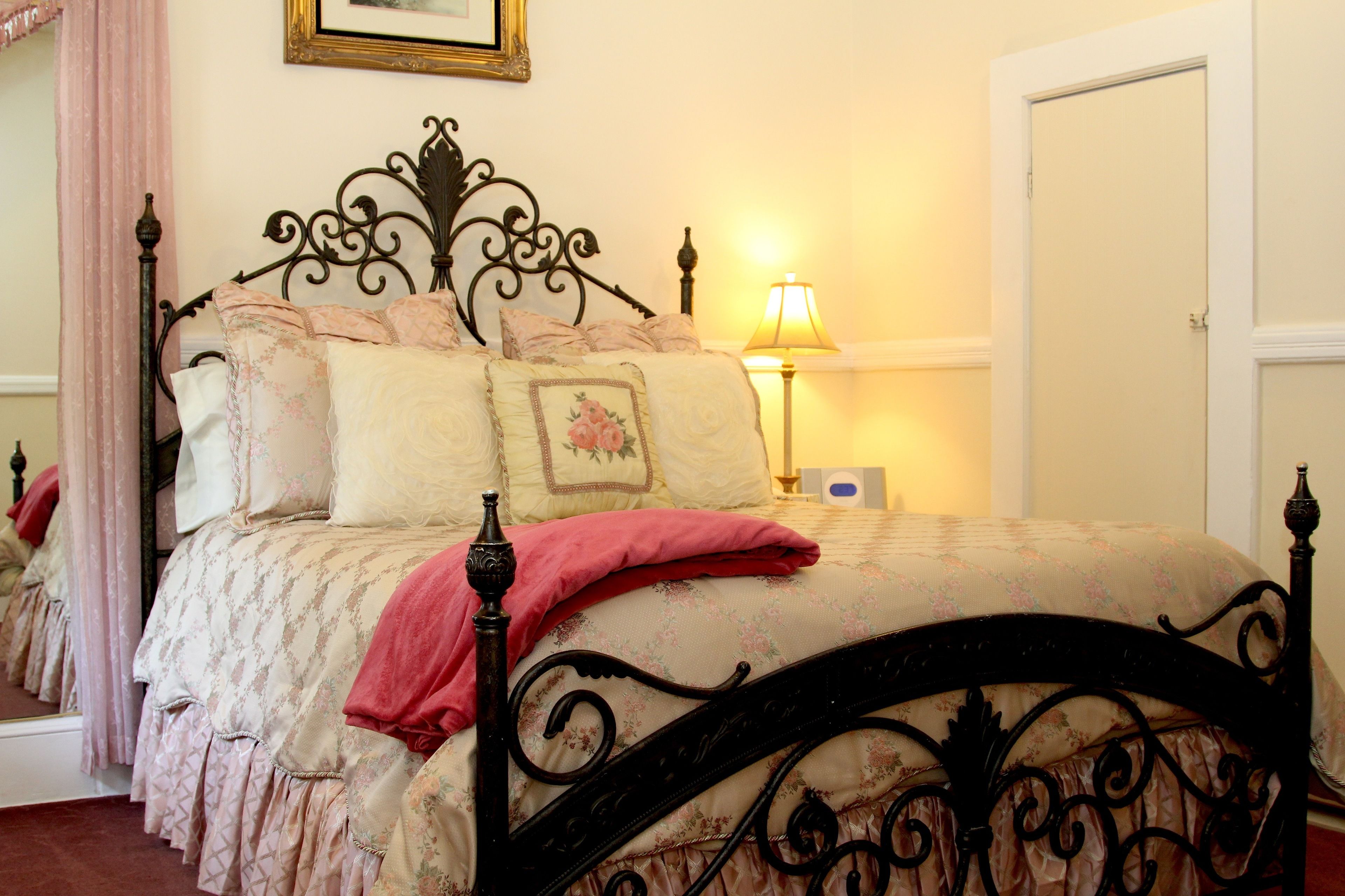 A neatly made bed with a floral bedspread and a red throw blanket, a bedside lamp, and a mirror reflecting the room.