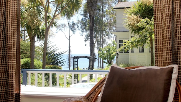 A wicker chair with a brown cushion, overlooking a balcony with a white railing, palm trees, and a view of the ocean. - Monarch Cove Inn (Capitola)