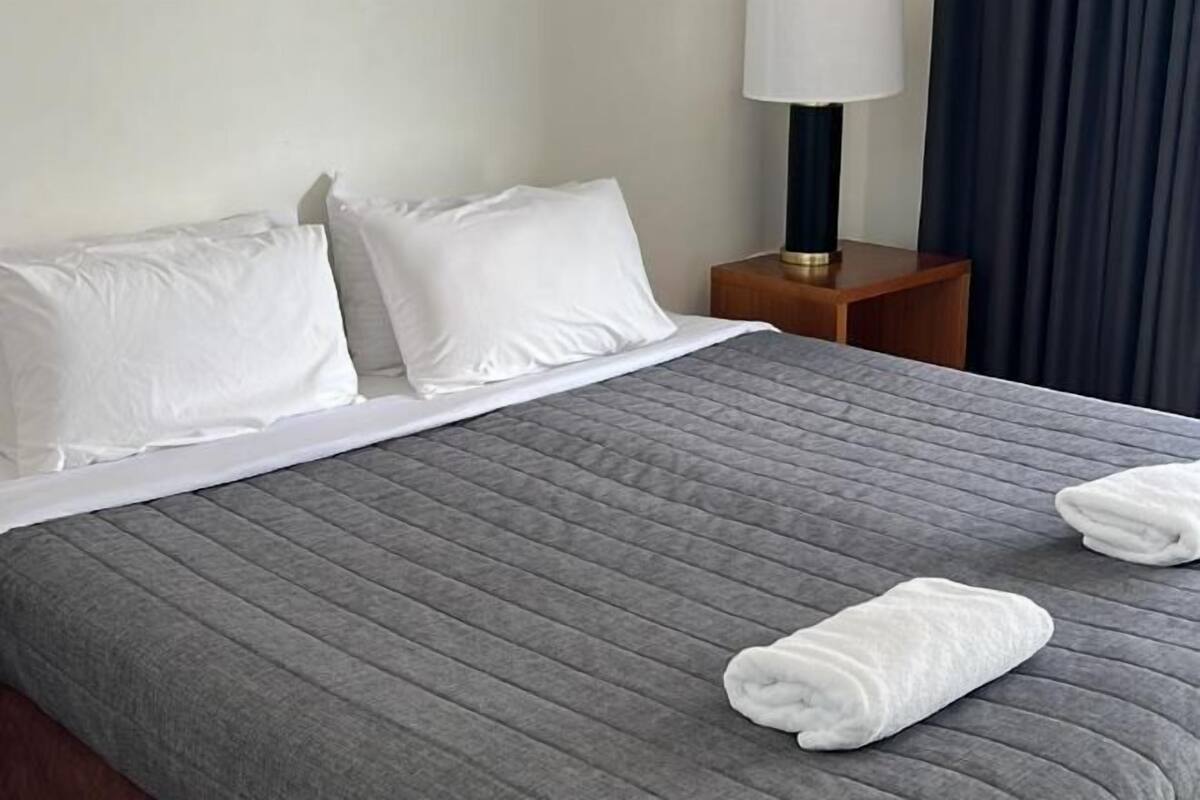 A neatly made bed with white pillows and a grey quilted bedspread. There are two folded white towels on the bed.