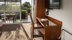 Habitación de hotel con un escritorio de madera, un televisor de pantalla plana montado en la pared, una puerta de cristal que da a un balcón con mesa y sillas, y suelo alfombrado.