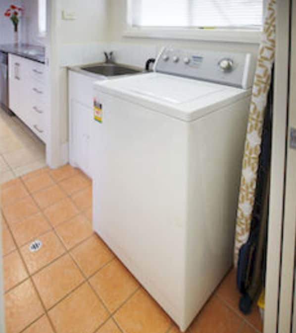 A compact, white washing machine in a small kitchen with a tiled floor and a window with blinds.