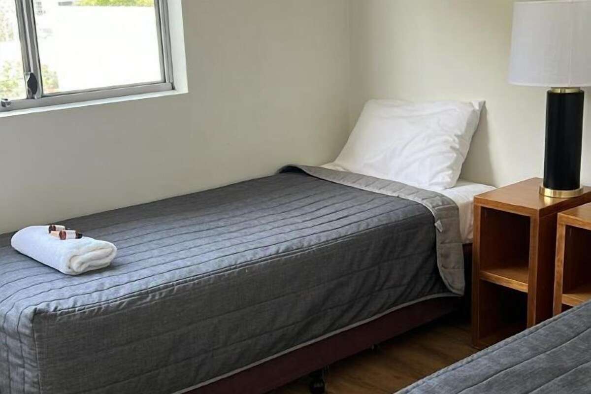 Un lit simple avec une couvre-lit grise, un oreiller blanc, une table de chevet en bois avec une lampe, et une fenêtre donnant sur l’extérieur.