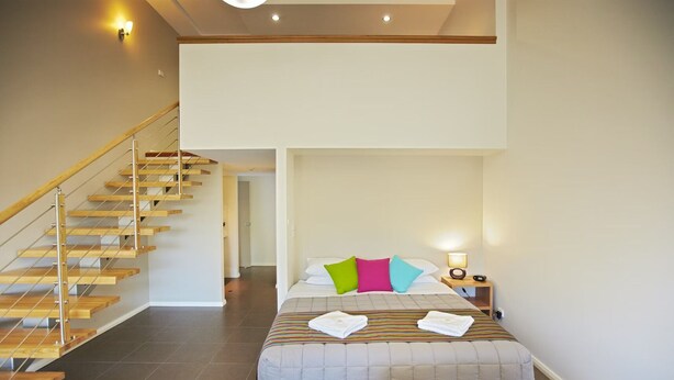 Un dormitorio con cama, almohadas coloridas, una mesita de noche con lámpara y una escalera de madera.