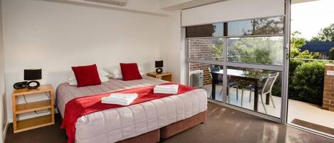 A modern hotel room with a large bed, a sliding glass door to the balcony, and a small table with chairs outside.