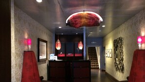 Um hall de entrada com balcão de recepção, duas poltronas vermelhas, um lustre grande e uma escadaria ao fundo.