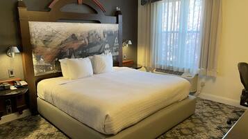 Quarto de hotel com uma cama grande, cabeceira com mural de paisagem urbana, mesa de cabeceira com abajur e janela com cortinas finas.