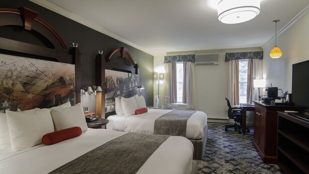 Quarto de hotel com duas camas, uma escrivaninha, uma cadeira, televisão e uma janela grande com cortinas.