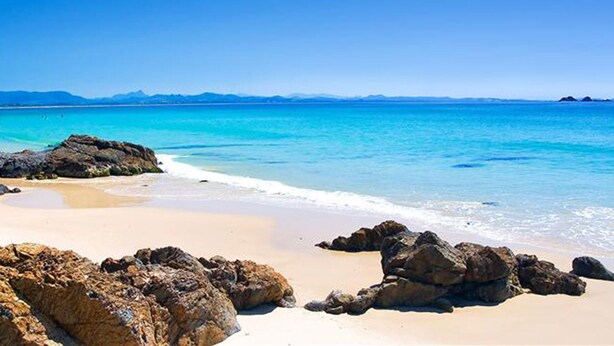 Une plage avec une eau turquoise et limpide, un rivage sablonneux et de gros rochers.