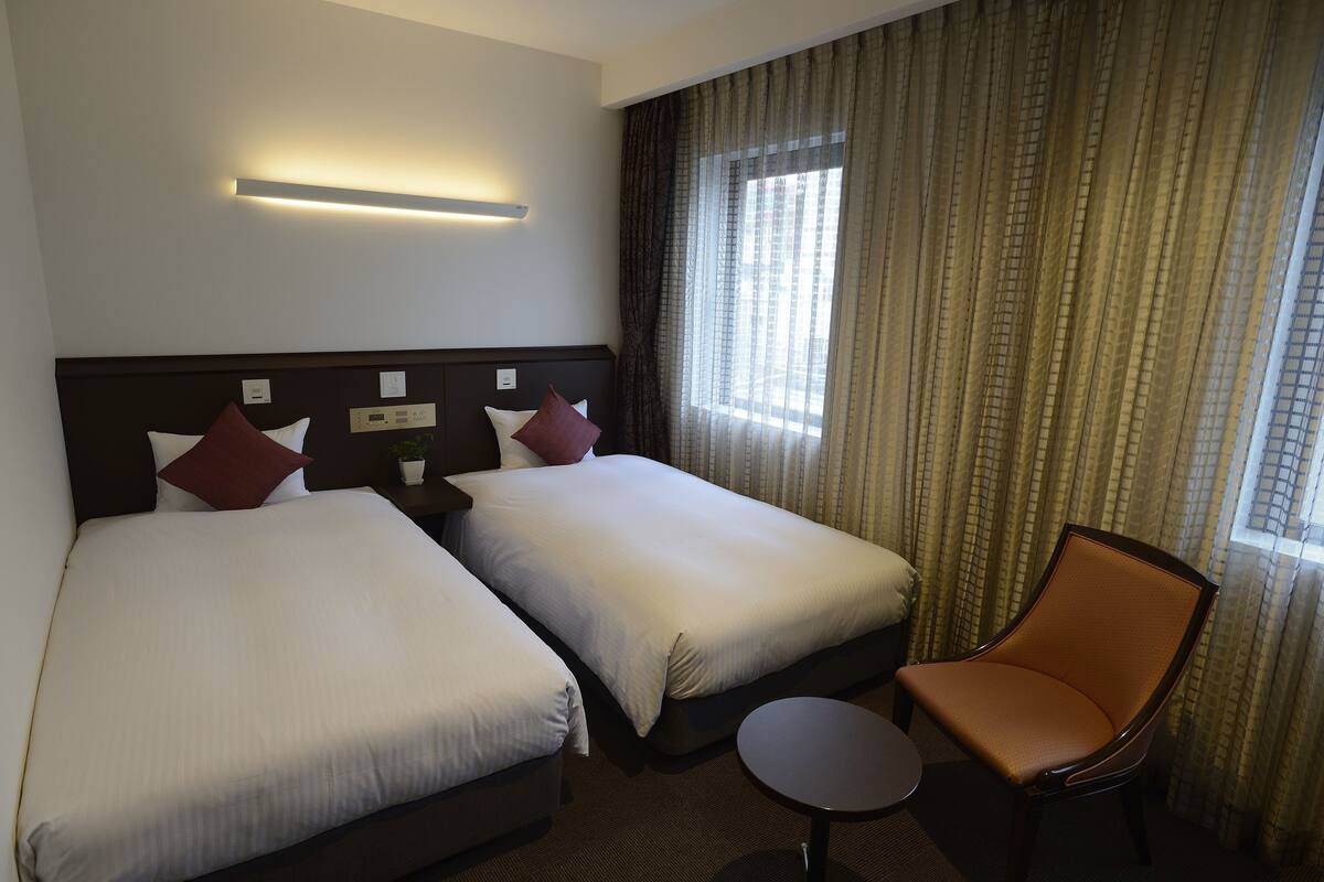 Habitación de hotel con dos camas, una silla, una mesita y una ventana con cortinas.