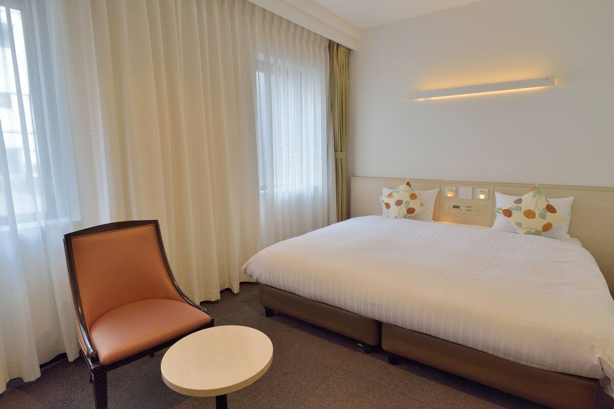 Una habitación de hotel con una cama, una silla, una mesita y un ventanal con cortinas.