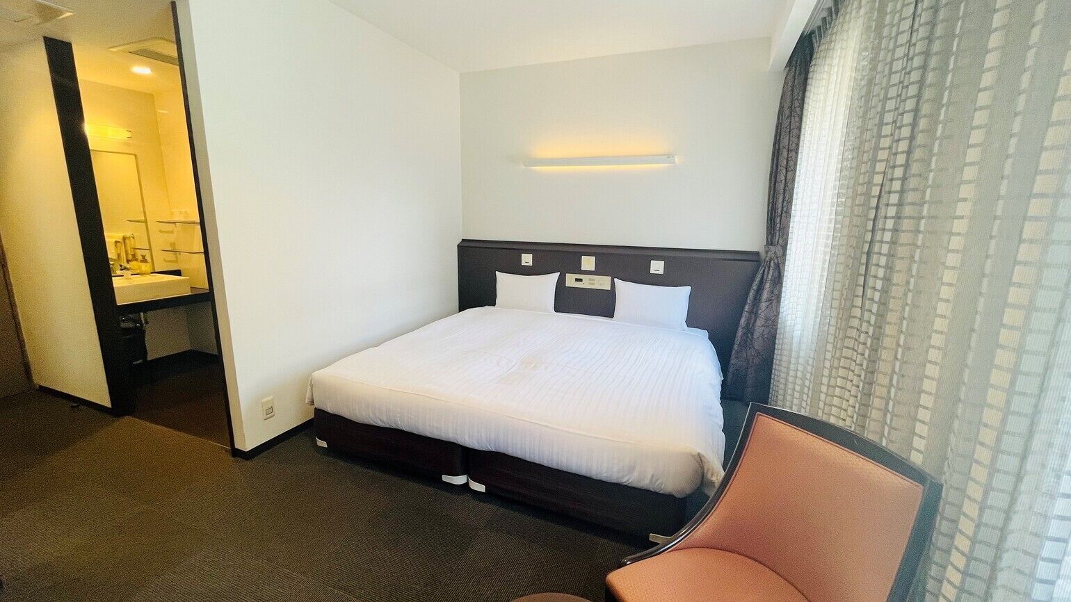 A hotel room with a bed, a chair, a bathroom with a mirror, and a window with curtains.
