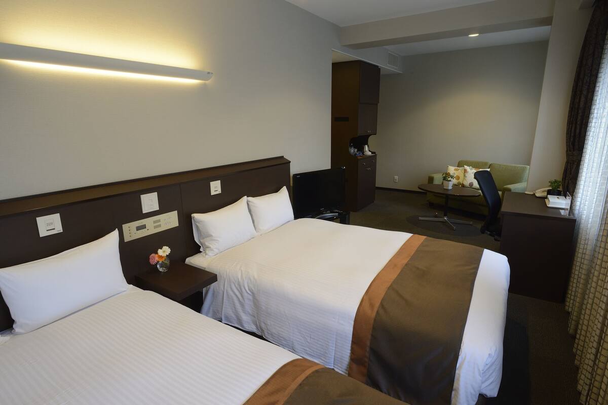Habitación de hotel con dos camas, un escritorio, una silla, una cafetera y una mesita con un jarrón de flores.