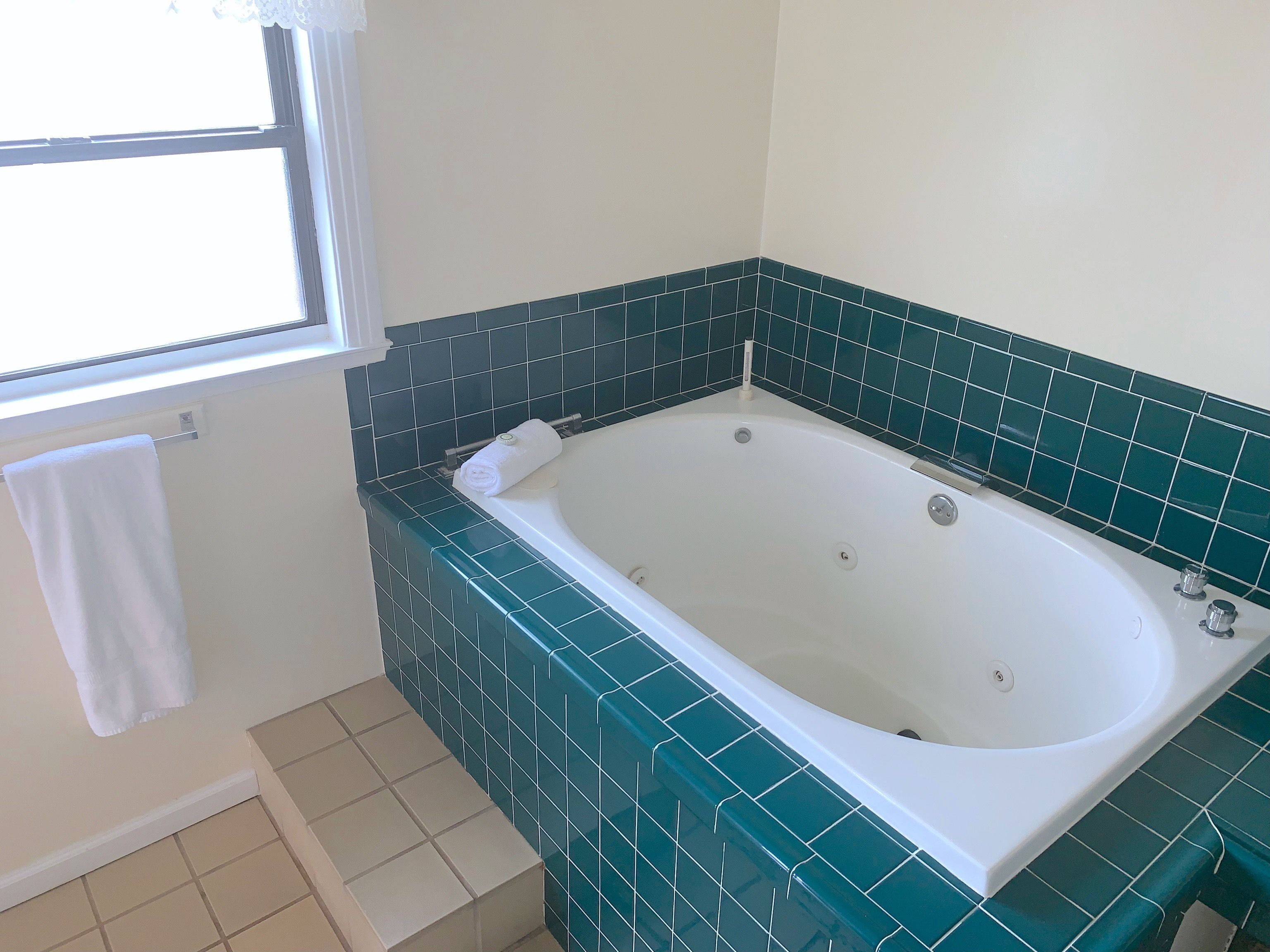 Une salle de bain avec une baignoire blanche, des murs recouverts de carreaux verts, une fenêtre avec une serviette blanche suspendue et un sol carrelé.