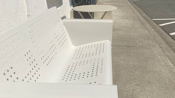 A row of white outdoor benches with perforated backs, positioned on a sidewalk next to a building with a white exterior.