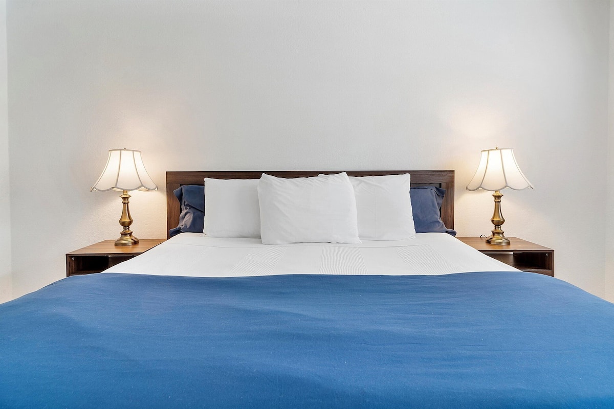A neatly made bed with white and blue bedding, flanked by two bedside tables with lamps, in a simple room with white walls.