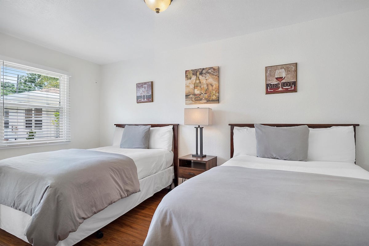 A hotel room with two beds, a nightstand with a lamp, and wall art depicting wine bottles and glasses.