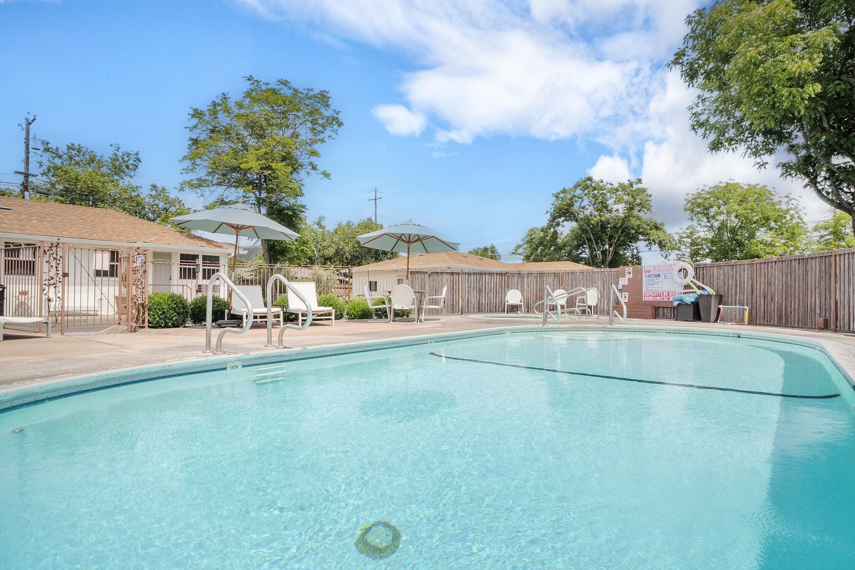 A swimming pool with lounge chairs and umbrellas, surrounded by a wooden fence and trees.