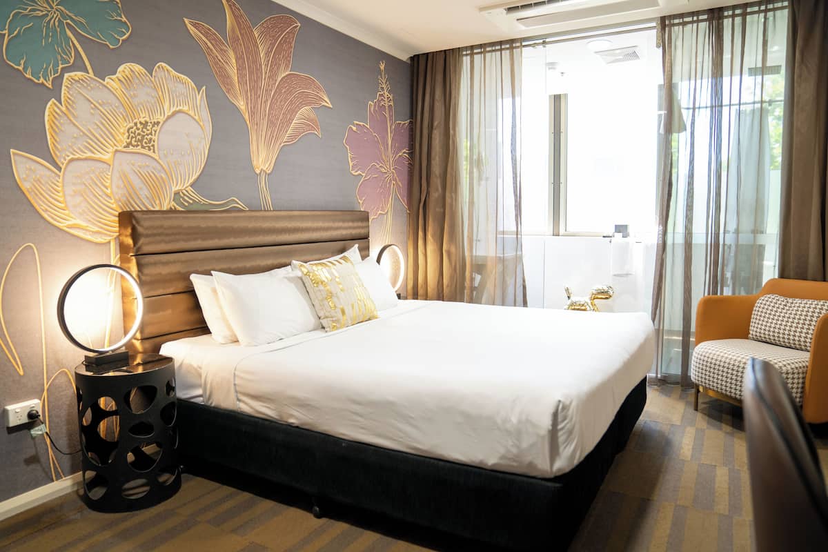 Habitación de hotel con una cama grande, una mesita de noche con lámpara, una silla y una pared decorada con motivos florales.