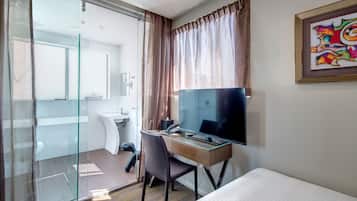 Habitación de hotel con cama, escritorio con computadora, televisor y baño visible a través de una puerta de vidrio.