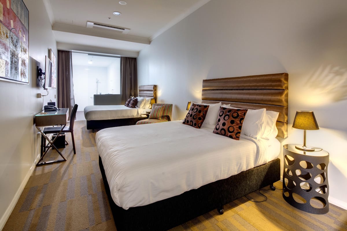 Habitación de hotel con una cama grande, un escritorio y una silla. Hay una ventana con cortinas, una lámpara y una obra de arte colgada en la pared.