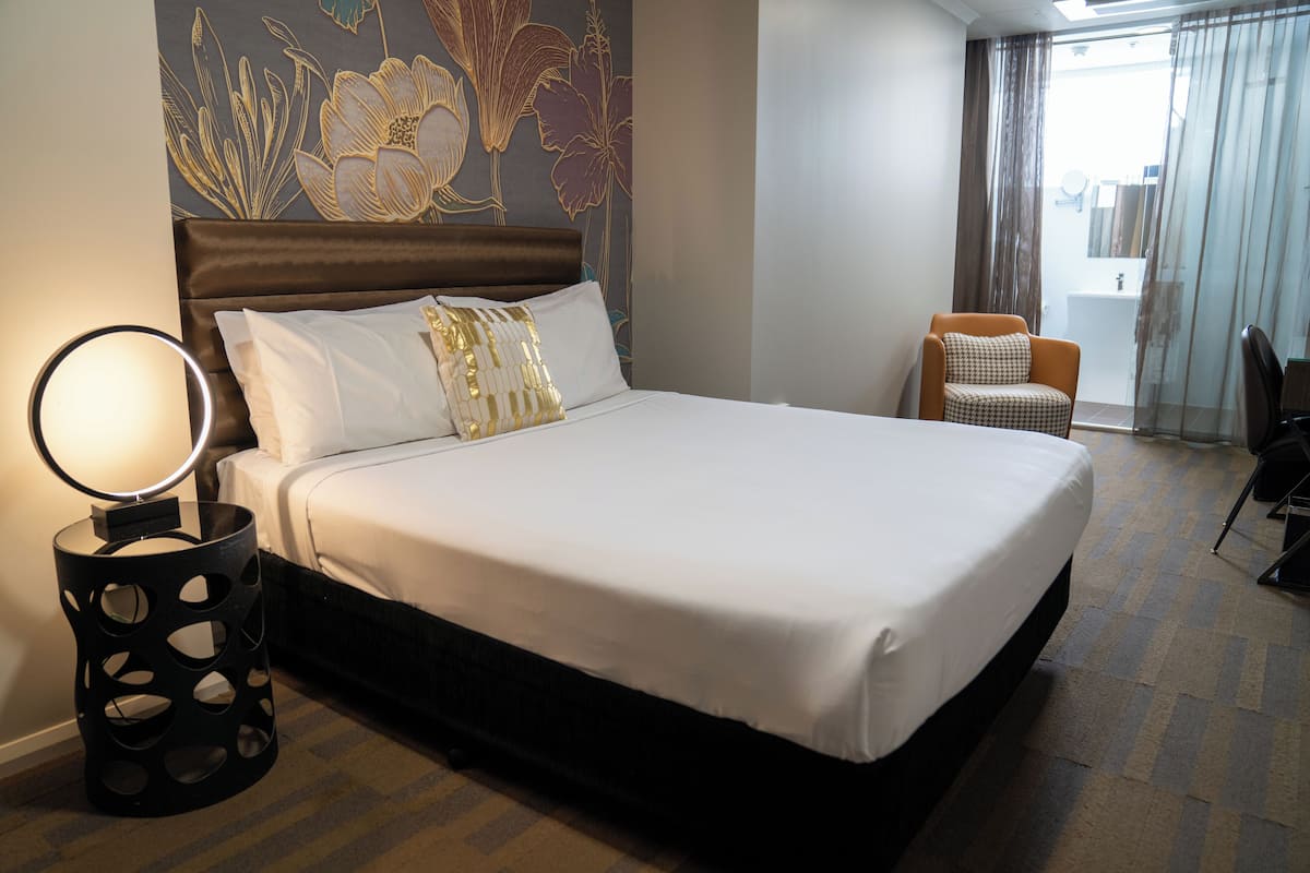 Habitación de hotel con una cama grande, una mesita de noche con lámpara, una silla y un baño visible a través de una puerta de cristal.