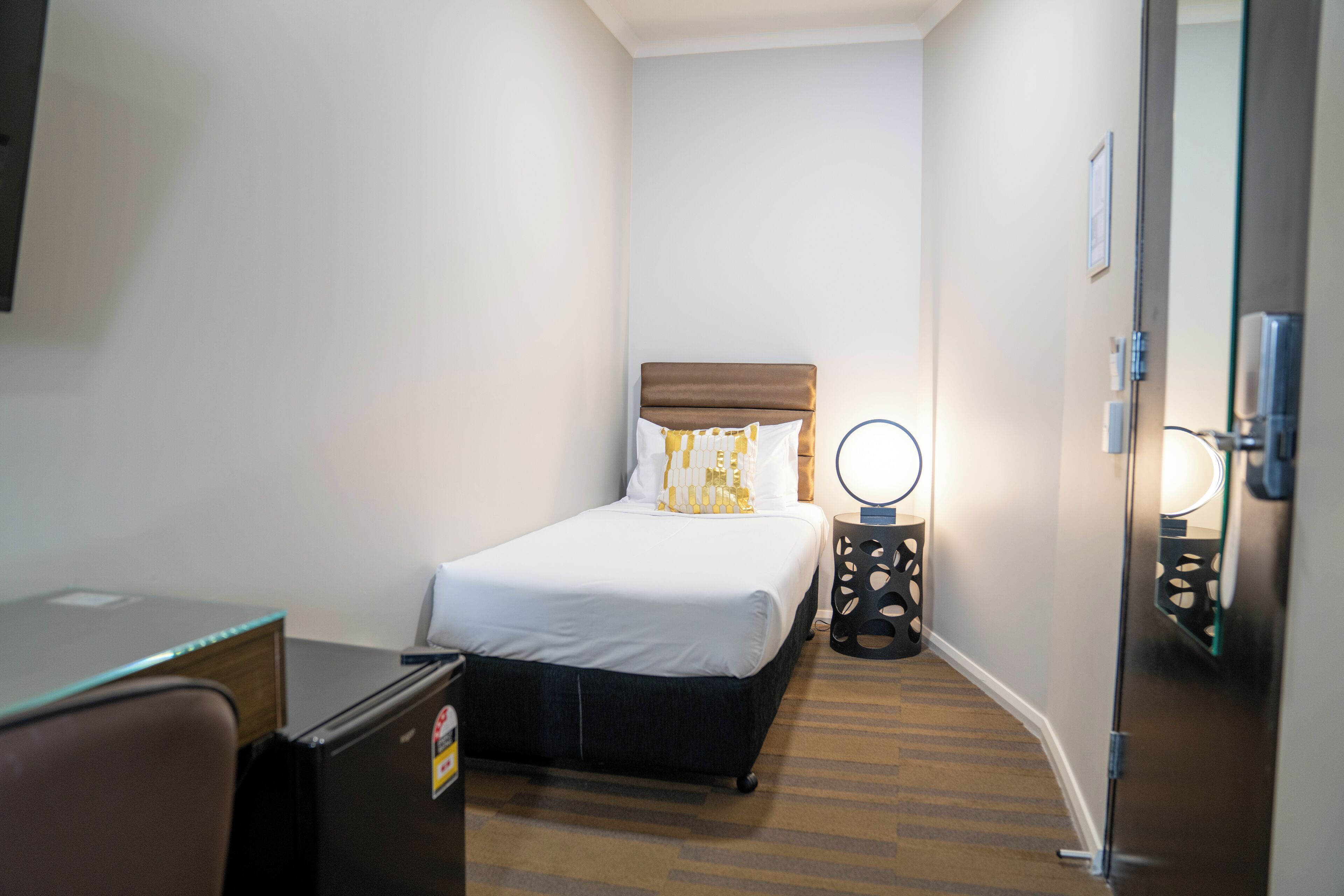 A compact hotel room with a single bed, a desk, and a small refrigerator.